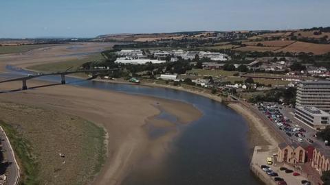 A drone shot of Barnstaple in Devon. It shows a big bridge crossing a river that is lacking water and mainly sand. There are houses and lots of cars parked along the quay side. There is also clear blue skies. 