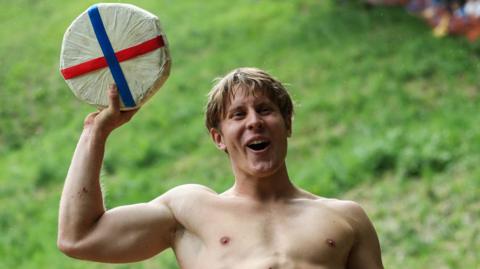 Tom Kopke, from Munich, holds a cheese wheel after winning the annual Cooper's Hill Cheese-Rolling  - he is bare-chested and has straight, fair hair. he has a smile on his face 
