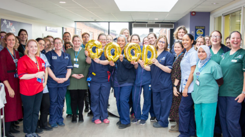 Salisbury District Hospital's maternity services employees stand in the hallway of the hospital dressed in their uniform, with three employees holding gold balloons that read 'GOOD'. They are all smiling at the camera. 