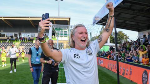 Robbie Savage holds his arms above his head, with a phone in one hand, as he smiles at fans while standing on the pitch