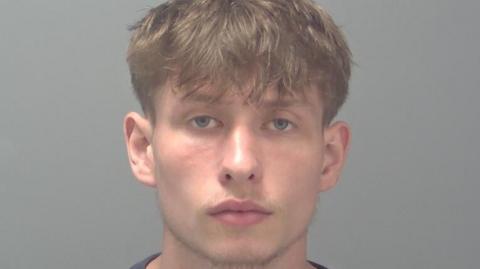 Jake McMillan is looking at the camera in a police custody photo. He has short light brown hair and blue or green eyes. He has a beard.