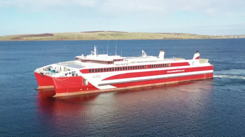 The MV Alfred sailing on the water. The boat is mainly red and white with a number of windows on the side. It is sitting on water which is dark blue in colour. It is a sunny day with blue sky above and a few white-grey clouds. A green hill is in the background.