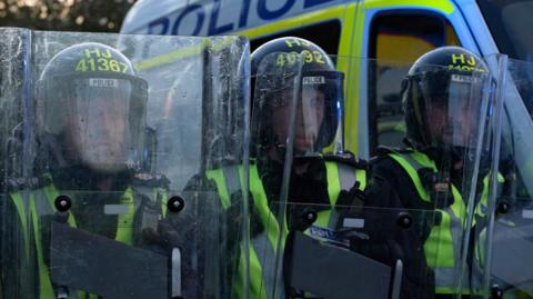 Police officers wearing helmets and high‑visibility vests stand close together holding clear riot shields, with a marked police van parked behind them.