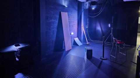 A room in a music venue. There is purple lighting in the room. A chair, a flooring tile, two brooms and a piece of wire mesh are in the room.