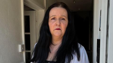 Nerys Walters stood in the door way of her home. She has dark black hair and dark red lipstick and is looking at the camera frowning.