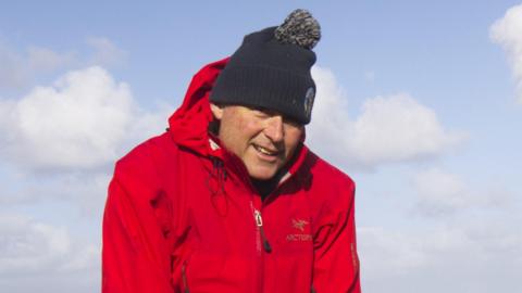 A man in a red waterproof coat, wooly hat and waterproof trousers stands in from of a body of water