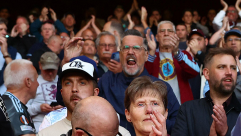 Aston Villa fans cheering in a stadium. They are wearing claret, blue and sky blue hates, jumpers and scarfs colours of the club