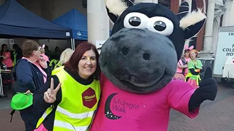 A woman wearing a yellow hi-vis jacket. She is cuddling a person dressed as a hippo mascot and wearing a pink Midnight Walk T-shirt.