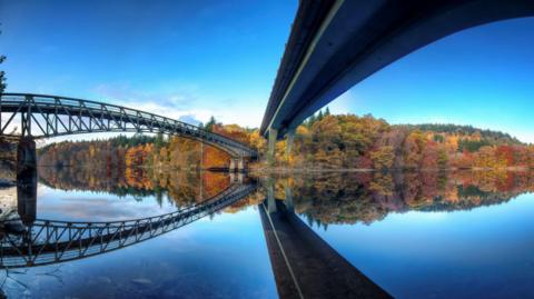 A pedestrian bridge, left, and a road bridge over Loch Faskally. The bridges are reflected in the mirror-like waters of the loch below. There are trees in autumn colours on the far shore.