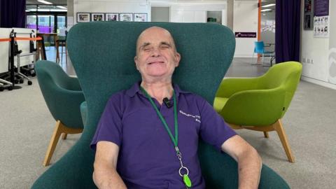 A man is wearing a purple t-shirt and a green lanyard around his neck. He is sitting in a green chair and smiling.