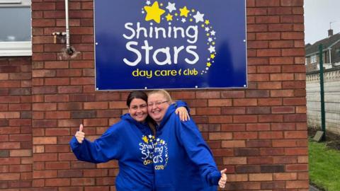 Two woman in blue hoodies smile and hug as they hold their thumbs up to the camera