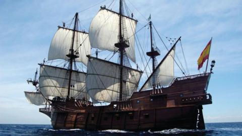 A huge 17th century galleon ship with a wooden hull and decks, six visible white sails and flying a Spanish flag whilst launched on the sea. 