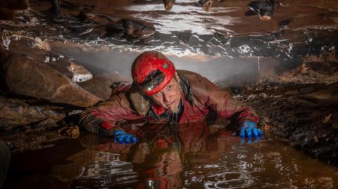 A caver from the exploratory team wearing waterproofs, blue gloves and a red helmet with a  light on on his front between a small gap of rock that sits above and below him. He has his hands on the wet ground in front of him and is looking at the camera.