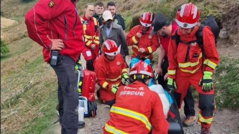 A group of seven emergency workers in red coats with reflective yellow bands, and two in a dark green uniform, along with a person in a baseball cap and grey coat, are stood around a person on a stretcher on a hillside path. Some are kneeling around a person on the stretcher. 