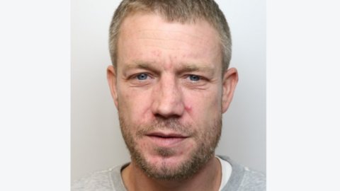 A mugshot of Steffon Wood wearing a grey crewneck sweatshirt. He has blue eyes and short brown hair which is turning slightly grey. He is standing against a grey background, looking straight at the camera with a blank expression.