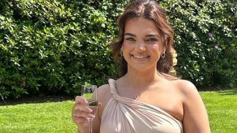 Amy White is pictured on a friend's wedding day. She is wearing a pink bridesmaid dress and is holding a champagne flute and a bouquet of gypsophila flowers. 