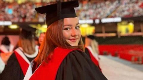 Nia Tyler has brown hair and is smiling at the camera. She is wearing a black graduation cap and gown, with a red sash.
