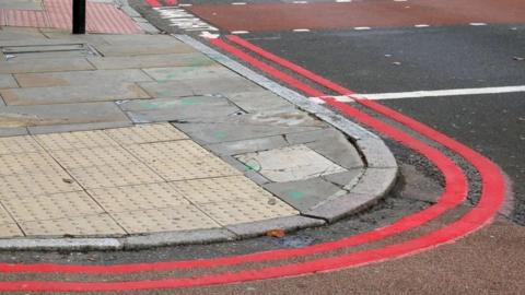 A close up ground-level shot of a dropped kerb at a junction with double red lines painted around it.