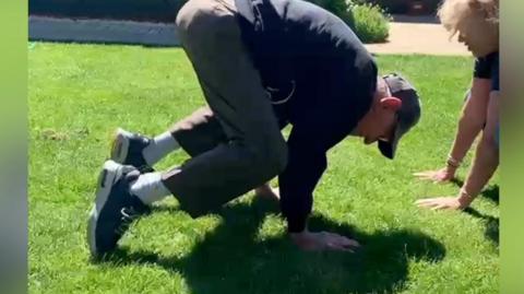 An elderly man has his hands on grass with bent knees facing the ground in a yoga position