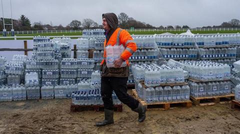 A person in an orange high vis jacket walking with water bottles under his arm. There are hundreds of bottles behind him.