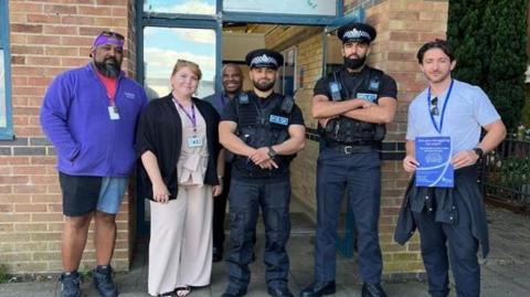 Five people smile at the camera as they stand in a line in front a building. Two are police officers dressed in uniform and three others who are wearing lanyards. 