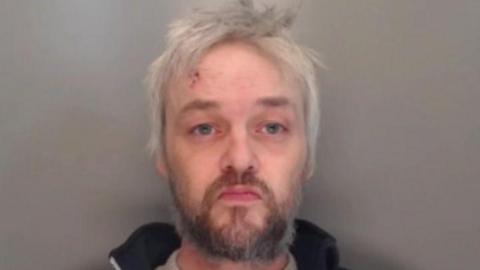 Mugshot of Tate. He has long messy straggly white hair and an untidy dark beard. He appears to have a cut on his forehead