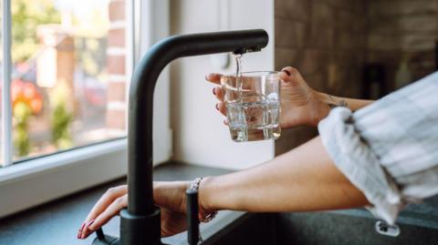 A black tap is releasing water into a small glass as someone with red nail varnish holds down the lever. They have a tattoo on their wrist and a rolled-up grey shirt.