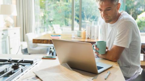 A man in a white t-shirt with stubble and short grey hair sits at a breakfast bar in a kitchen. He is looking into an open laptop and holding a green mug. In the background we can see a dining table, furniture and double doors that appear to open out into the home's garden. 