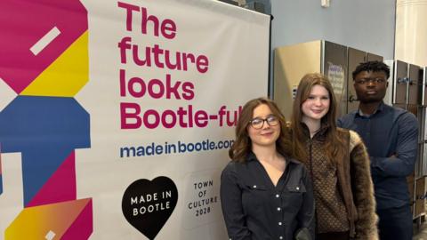 Three students - two girls one boy - stand in front of a sign at Hugh Baird college, Bootle which says" The Future is Bootle - Full"
