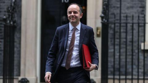 James Murray, a white man with dark hair, leaving No 10 wearing a suit, carrying a red folder.