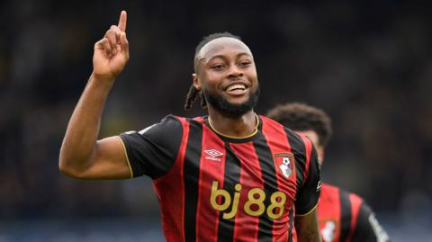 Antoine Semenyo, wearing a Bournemouth home shirt with red and black vertical stripes and gold sponsor's logo, raising his right forearm in the air and points with his index finger to celebrate a goal against Leeds in the Premier League
