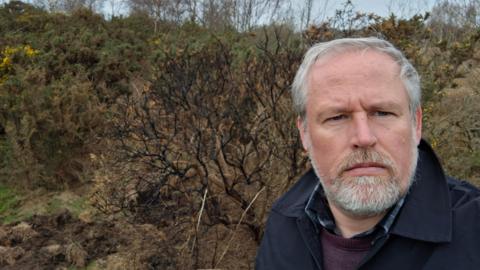 Durham County Councillor Mark Wilkes. He has short white hair and a trimmed beard. He has blue eyes and is wearing a blue coat, over a purple jumper. He is looking at the camera with a serious expression. Behind him are some gorse bushes.