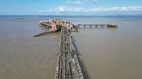 Birnbeck Pier which has fallen into disrepair. The wooden platform bridge is rotten and the planks are broken. The sea is brown and and the sky is blue with some white clouds in the distance.