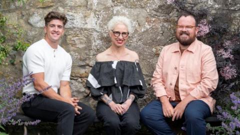 Danny Campbell, Anna Campbell-Jones and Banjo Beale sit on a bench in front of a stone wall in a garden. There are plants and flowers climbing the wall and surrounding the judges.