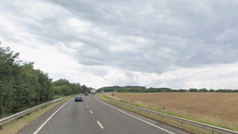 The A417 near Birdlip with cars driving in the distance. There is a wheat field to the right of the road and grey sky overhead.