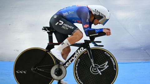 William Salter riding on a cycling velodrome track at the National Track Championships wearing a blue long sleeved skinsuit with black shorts covered in advertising branding. He is wearing a white helmet with the visor down, while his bike is black with black track wheels