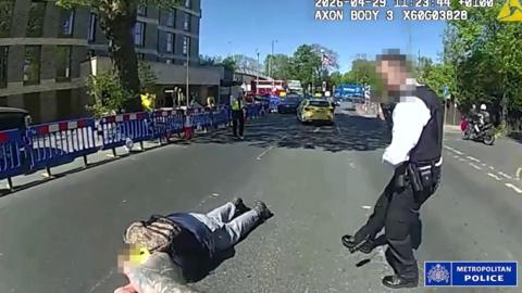 A bodycam image - a man lies face down on the ground in the middle of the road in an urban area as a police officer stands over him.