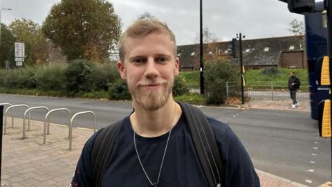 Tom has short blonde hair and blonde facial hair. He is standing in front of a road.