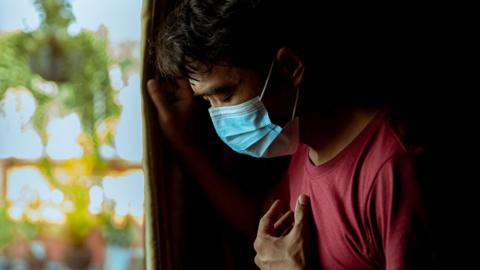 An asian man of unknown age stands by a window in a red tshirt and wearing a blue face mask. HIs head is bowed and his eyes are closed as if sad or contemplative and he holds a hand to his chest. 