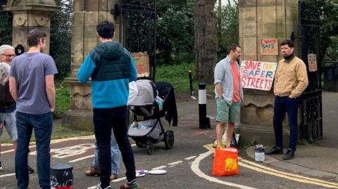 Several people standing outside park next to placard that says 'save our streets'