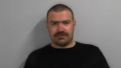 Head and shoulders police mugshot of Oliver James Anglesmith who has close-cropped dark hair, and stubbly facial hair. He is wearing a black T-shirt.