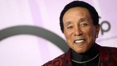 Motown legend Smokey Robinson is seen photographed on a red carpet. The close-up shot shows him in a black shirt and red, floral suit jacket. He is wearing two silver chains and smiling. 