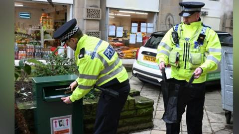 Two police officers in uniform using a knife disposal bin in Huddersfield town centre. One is putting a knife in the bin and the other is holding a bag open