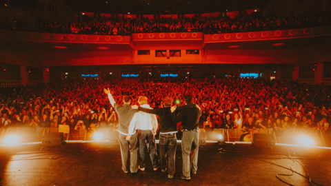 A picture of the band, with their backs to the camera, thanking fans for their support. Bright lights are in front of them and the fans are bathed in bright orange light. 