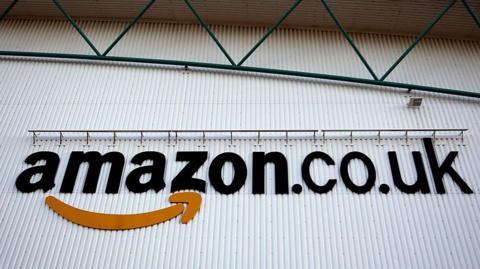 A logo sits on a wall outside the Amazon Marston Gate fulfilment centre, the building is white with green metal above. 