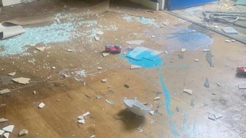 A wooden floor covered in broken glass and blue paint. There are also various bits of broken materials on the floor. There is a dark blue wall in the background.
