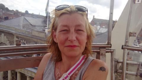 Ms Sturgess has dyed blonde hair pushed back by sunglasses and is wearing a grey vest top with a pink and white bag strap across her chest.
