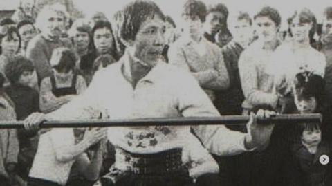 A black and white photo of a man holding a long stick in front of a crowd. 