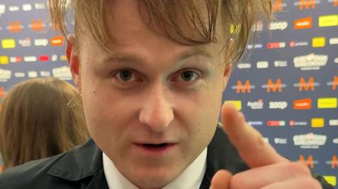 A man with short brown hair and wispy fringe points to the camera