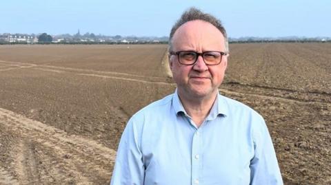A man with greying hair, wearing glasses and a light blue shirt, is standing on a farmland. 